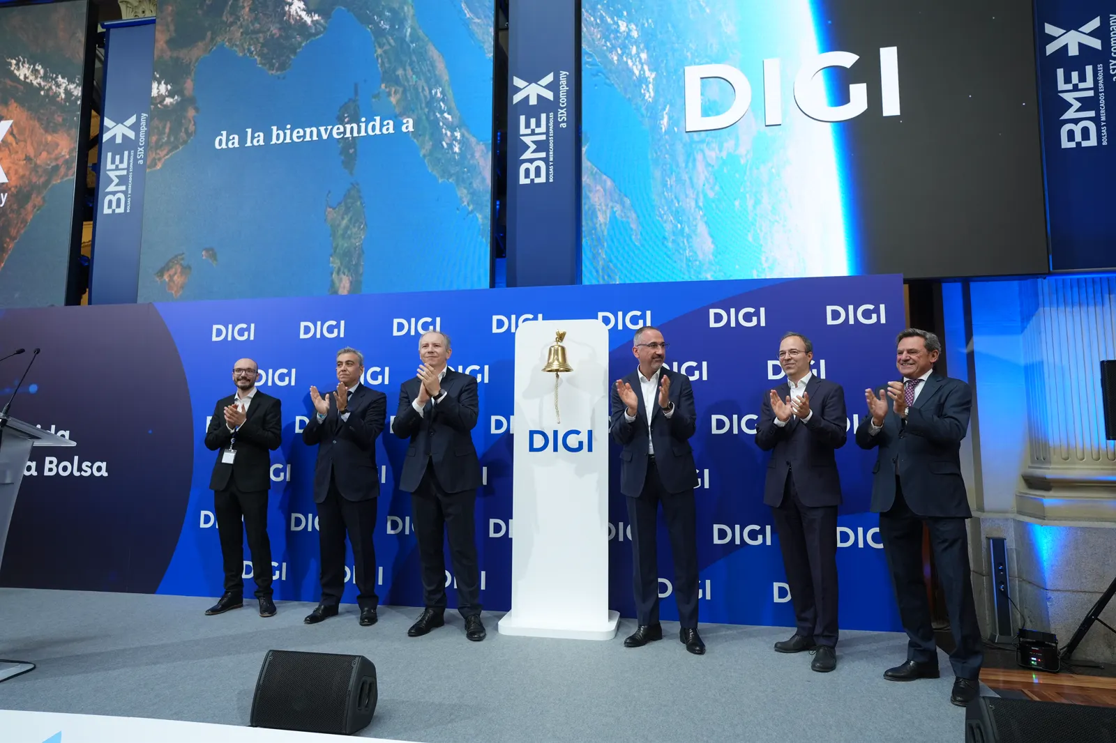 Seis hombres trajeados aplauden alrededor de un pedestal blanco con una campana dorada y el logotipo de DIGI, en un escenario rodeado de pantallas con los logotipos de DIGI y BME.