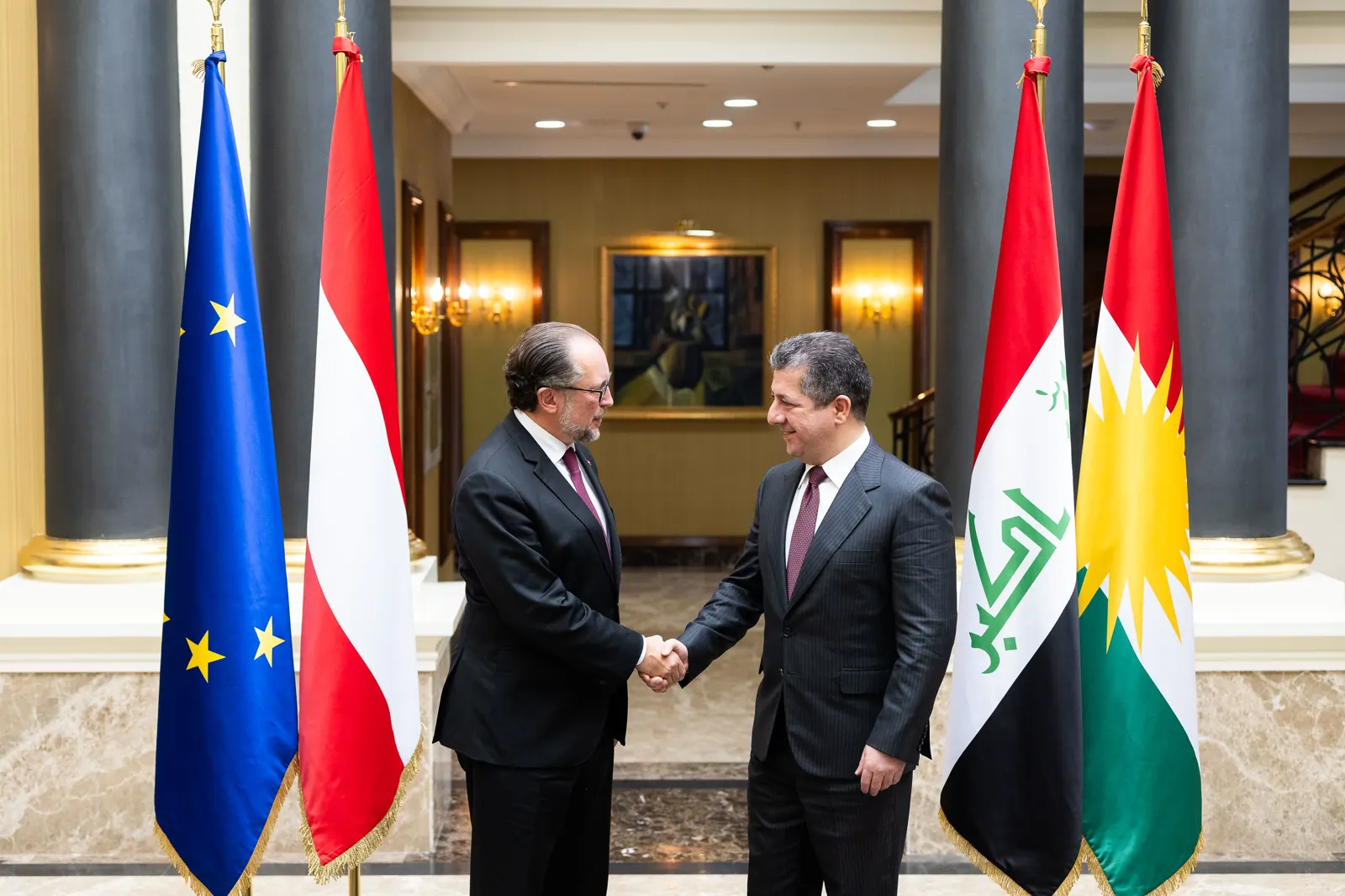 Dos hombres trajeados se estrechan la mano en un salón institucional, entre banderas de la Unión Europea, Austria, Irak y el Kurdistán.