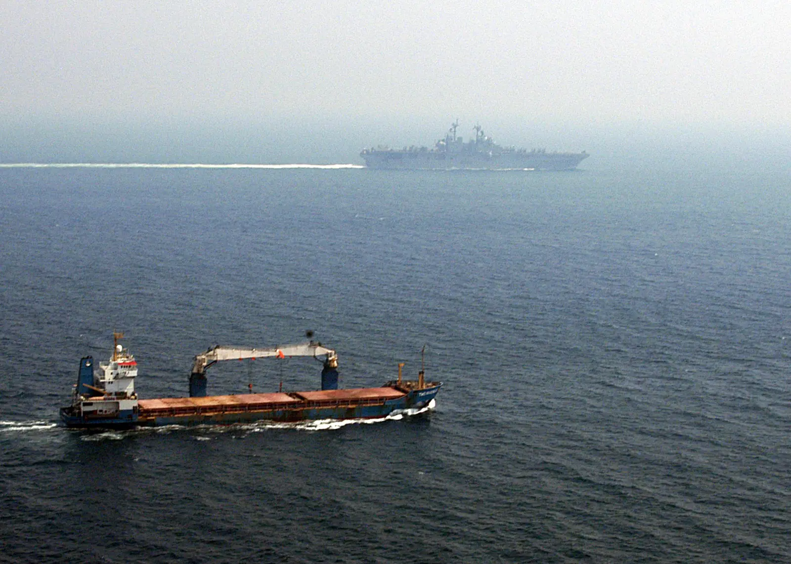 Un carguero azul con grúa de cubierta navega en primer plano por un mar en calma; al fondo, entre la calima, se recorta la silueta gris de un gran buque de guerra.