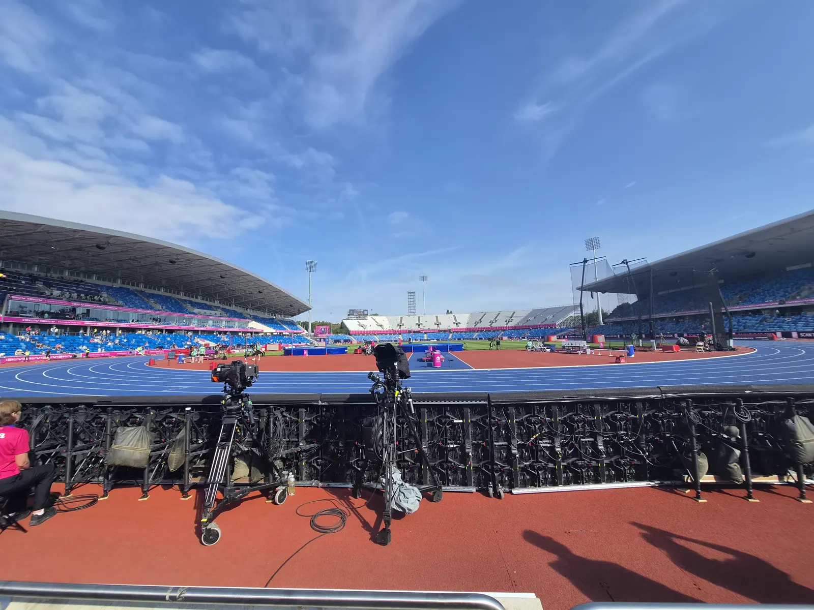 Pista de tartán azul y gradas del Alexander Stadium de Birmingham vistas desde la zona de prensa, con dos cámaras de televisión sobre trípode en primer plano y cielo despejado.