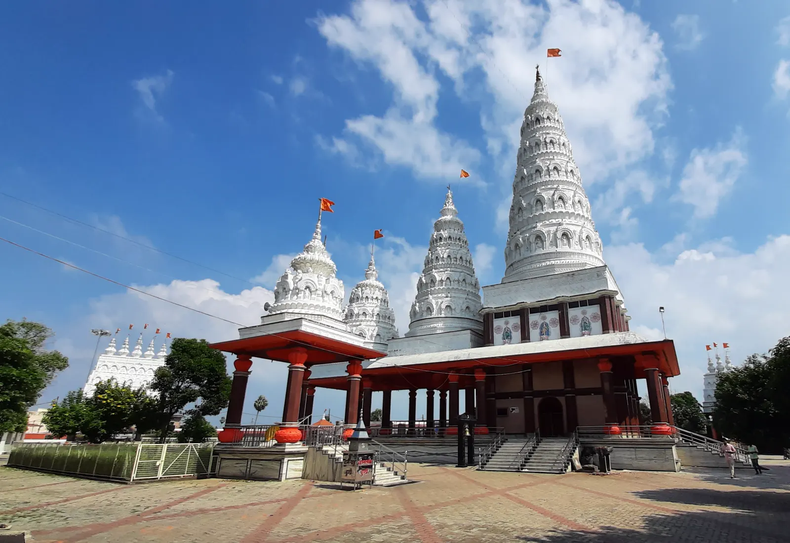 Templo hindú de fachada blanca con cuatro torres cónicas escalonadas rematadas por banderines naranjas, sobre un cielo azul con nubes. Delante, un pórtico de columnas rojas y una escalinata de piedra que baja a una explanada enlosada, vacía de gente.