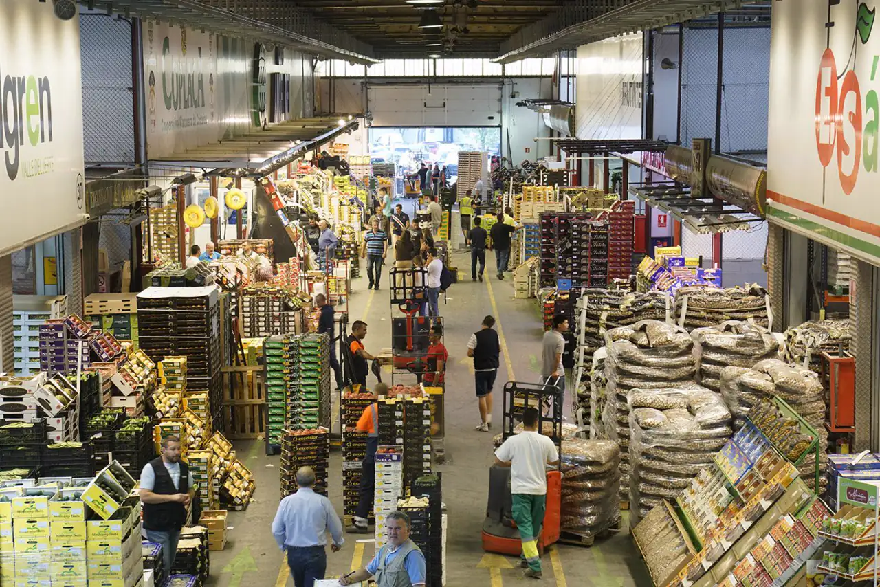 Pasillo central de una nave de Mercamadrid con puestos mayoristas a ambos lados, cajas de fruta y verdura apiladas hasta la altura de las personas y una veintena de trabajadores moviendo palés con transpaletas.
