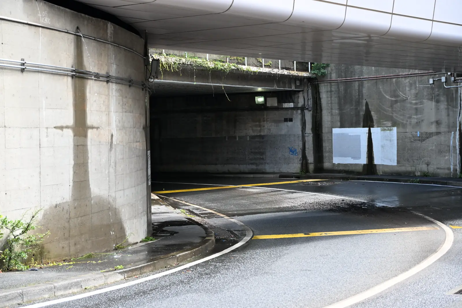 Boca de un paso inferior de carretera bajo una estructura de hormigón: la calzada, mojada, conserva restos de barro y sedimento junto al bordillo, y por los muros de contención escurren regueros de agua. No se ve a nadie ni hay vehículos.