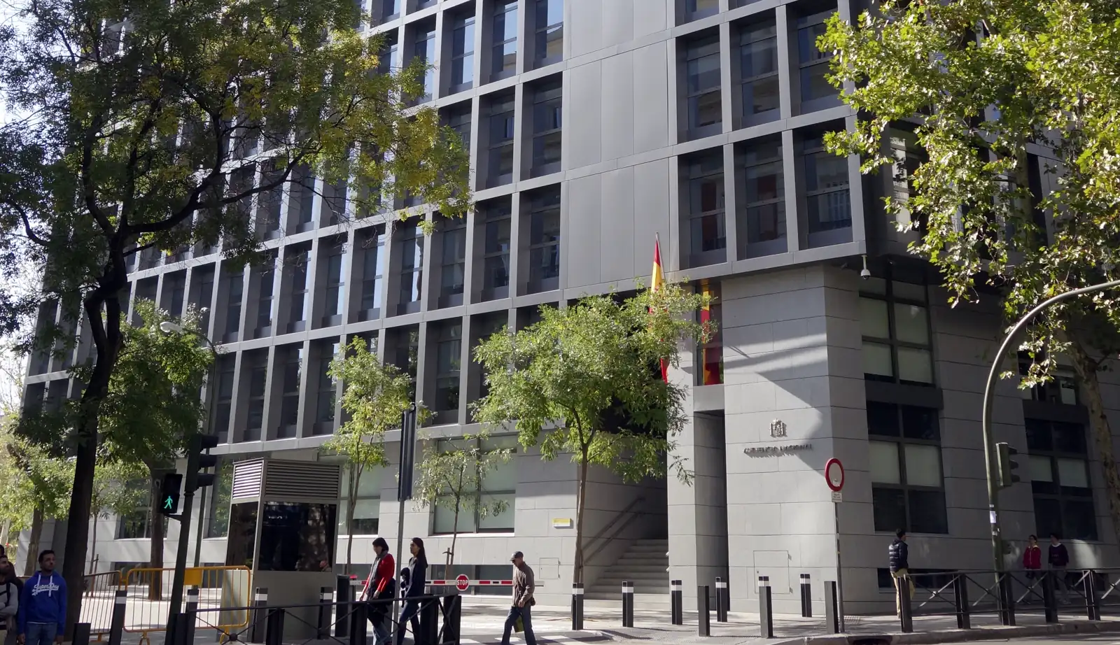 Fachada acristalada y gris de un edificio judicial de varias plantas en una calle arbolada de Madrid. Junto a la entrada ondea una bandera de España y en la esquina se lee, bajo el escudo, el rótulo «Audiencia Nacional». Varios peatones cruzan la acera en primer plano.