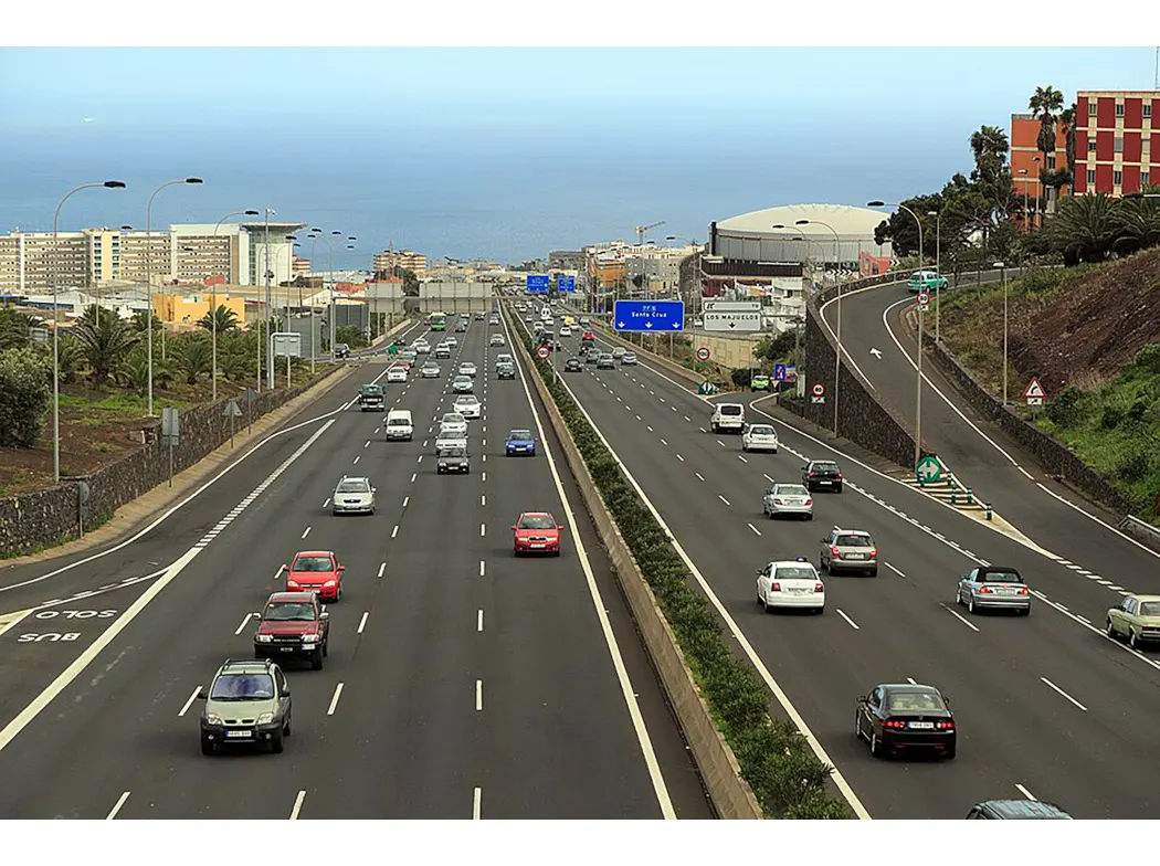 Vista elevada de una autovía de varios carriles por sentido con tráfico fluido de turismos; al fondo, bloques de viviendas, palmeras y el mar.