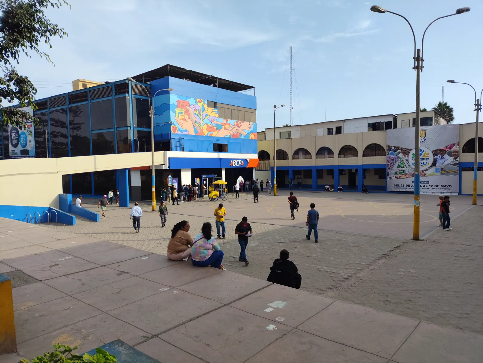 Plaza pavimentada de Villa El Salvador con vecinos caminando y sentados, farolas y edificios bajos de fachada azul y crema, uno de ellos con un mural de colores y una sucursal bancaria en los soportales.