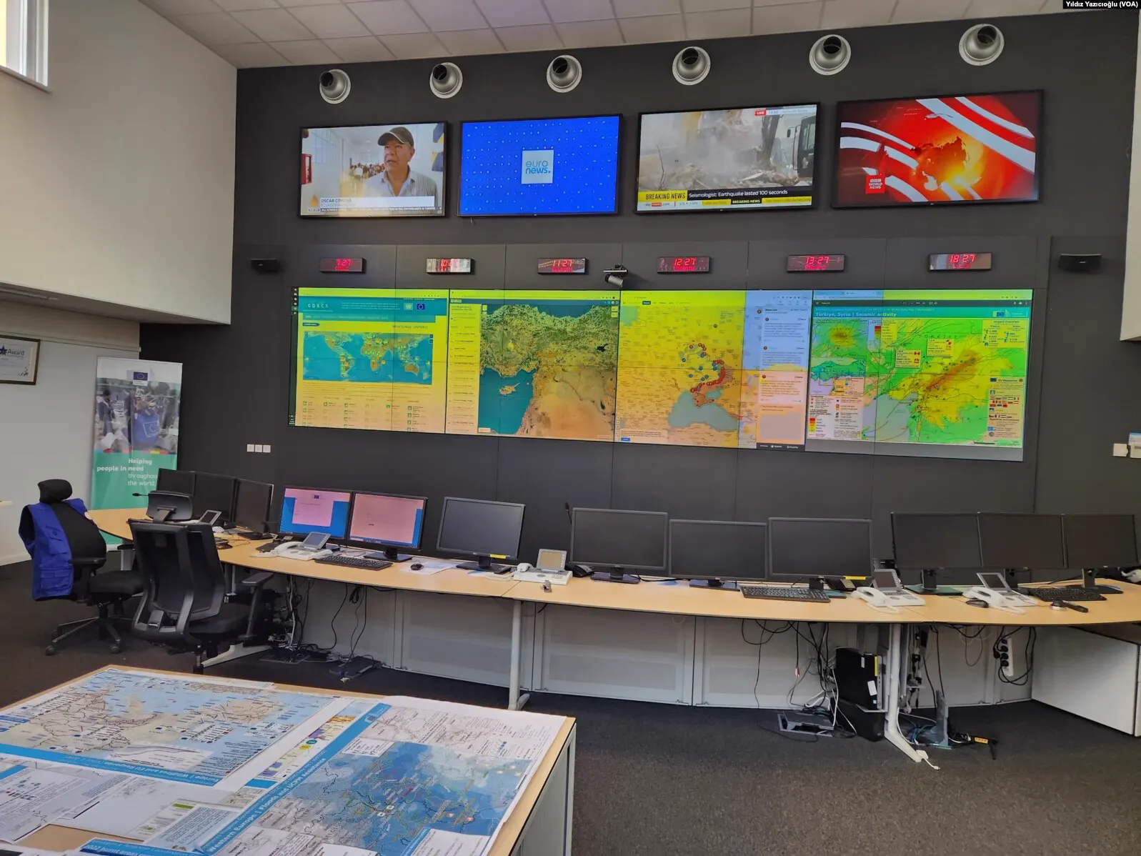 Sala de control del Centro de Coordinación de la Respuesta a Emergencias de la Comisión Europea: una mesa curva con una hilera de monitores apagados y, en la pared, una pantalla gigante con mapas de riesgo, cuatro televisores con canales de noticias y seis relojes con husos horarios distintos.