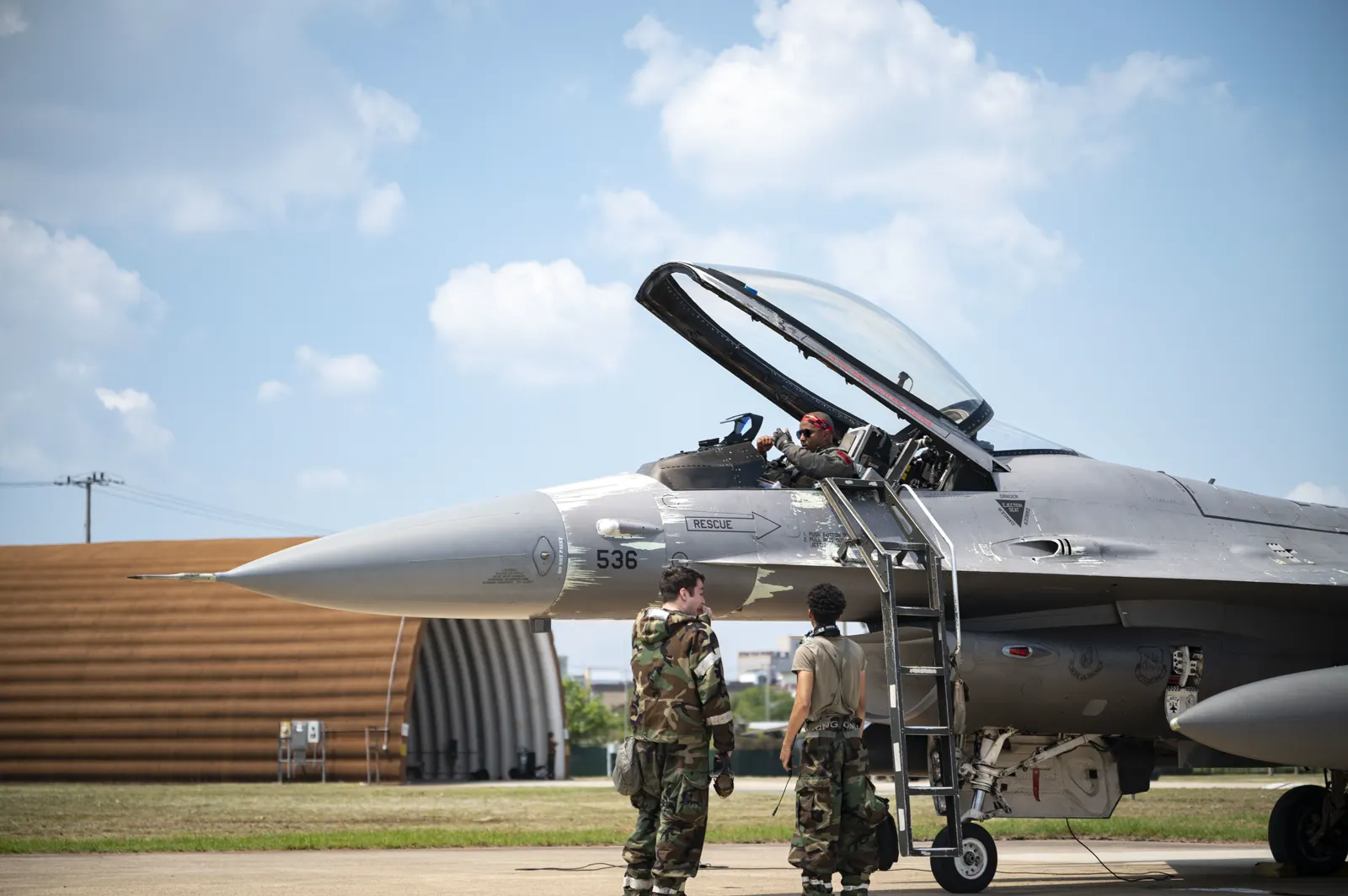 Un caza F-16 estadounidense estacionado en la pista de una base aérea con la cabina abierta y el piloto dentro; dos militares con uniforme de camuflaje esperan junto a la escalerilla y al fondo se ve un refugio de aeronaves.