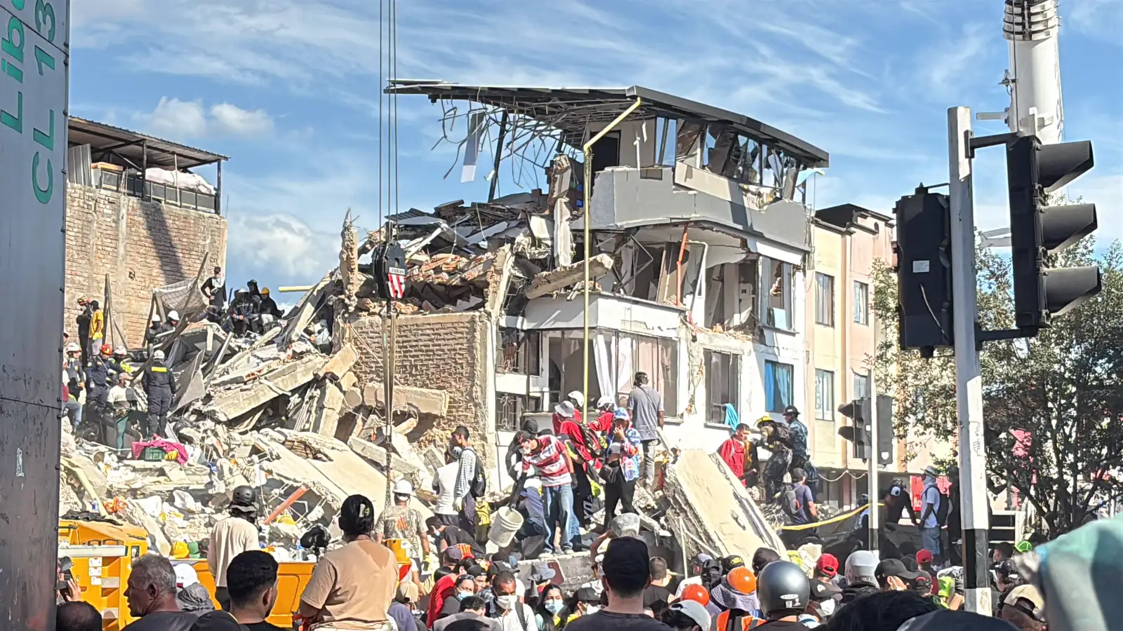 Edificio de varias plantas derrumbado sobre una calle de Pereira (Colombia). Sobre la montaña de escombros trabajan equipos de rescate con casco y chaleco reflectante, y decenas de personas se agolpan en la acera junto a un semáforo, mirando hacia las ruinas.