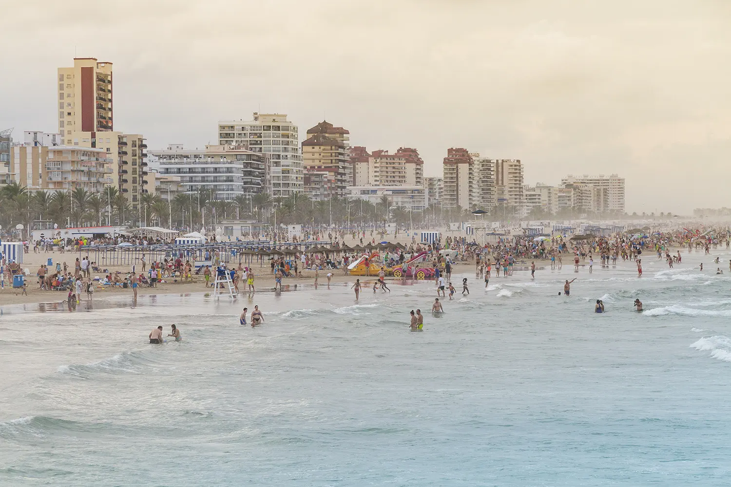 Vista de la playa de Gandía llena de bañistas y sombrillas, con la línea de bloques de apartamentos al fondo y el mar en primer término bajo un cielo blanquecino.