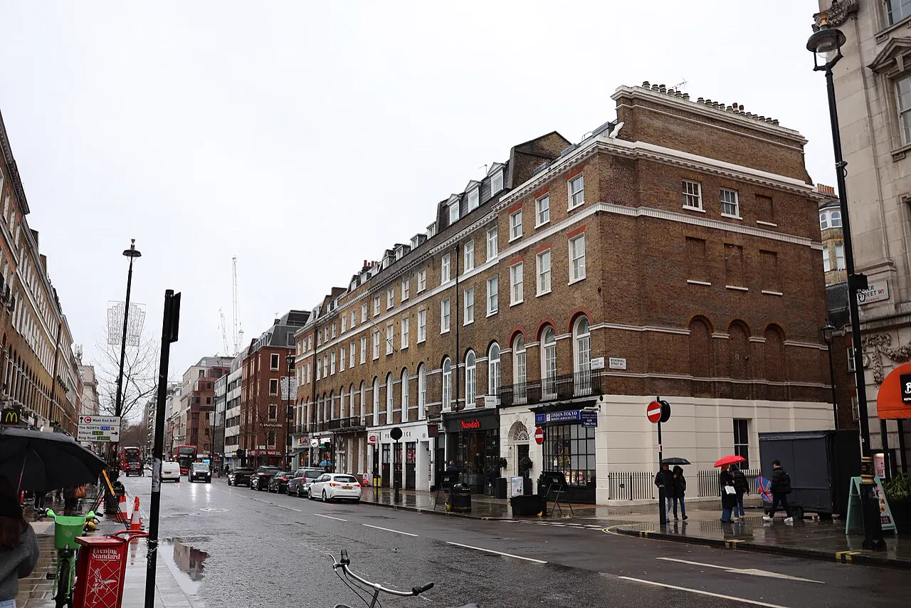 Vista de Baker Street en un día de lluvia: edificios de ladrillo de cuatro alturas con bajos comerciales, coches aparcados y peatones con paraguas en la acera.