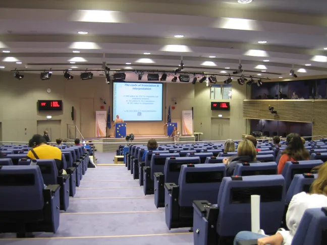 Interior de la sala de prensa de la Comisión Europea: filas de butacas azules en pendiente, casi todas vacías, con pupitres abatibles, orientadas hacia un estrado con atril, dos banderas de la Unión Europea y una pantalla de proyección. Del techo cuelgan focos y paneles luminosos; a la derecha, las cabinas de interpretación.