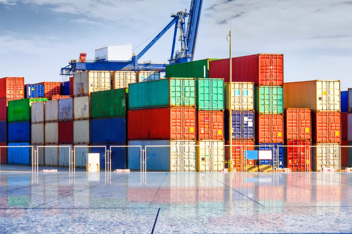 Contenedores de carga de colores apilados en varias alturas en la explanada de una terminal portuaria, con una grúa portuaria azul al fondo y el suelo mojado reflejando las cajas.