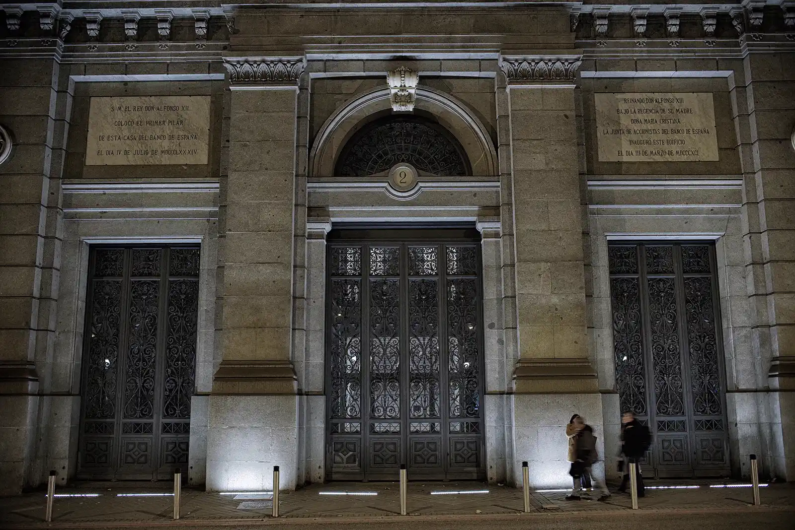 Fachada de piedra del Banco de España iluminada de noche: tres puertas de hierro forjado bajo arcos, dos lápidas conmemorativas grabadas en el muro y dos viandantes cruzando la acera.