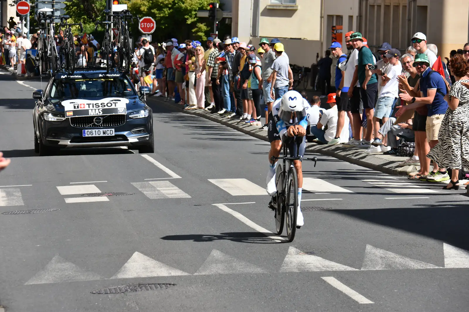Un ciclista con el maillot azul y blanco del Movistar Team pedalea en posición aerodinámica sobre una bicicleta de contrarreloj por una calle urbana, entre el público, seguido por el coche del equipo.
