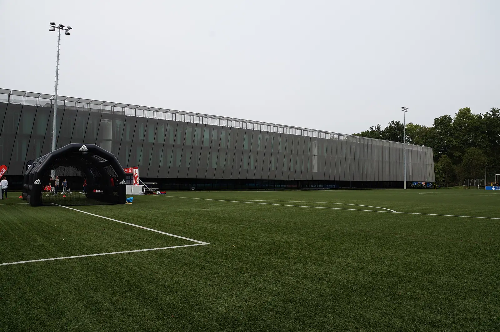 Edificio alargado y acristalado de la sede de la FIFA en Zúrich visto desde un campo de fútbol de césped, con una torre de focos, una carpa hinchable negra y unas pocas personas junto a la banda