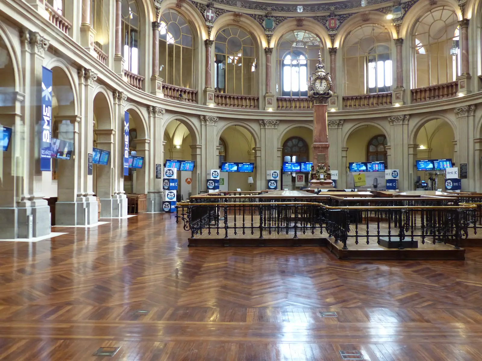 Sala circular del Palacio de la Bolsa de Madrid, con suelo de parqué, barandilla de hierro forjado alrededor del corro, un reloj de bronce sobre columna de mármol y pantallas azules con cotizaciones bajo los arcos.