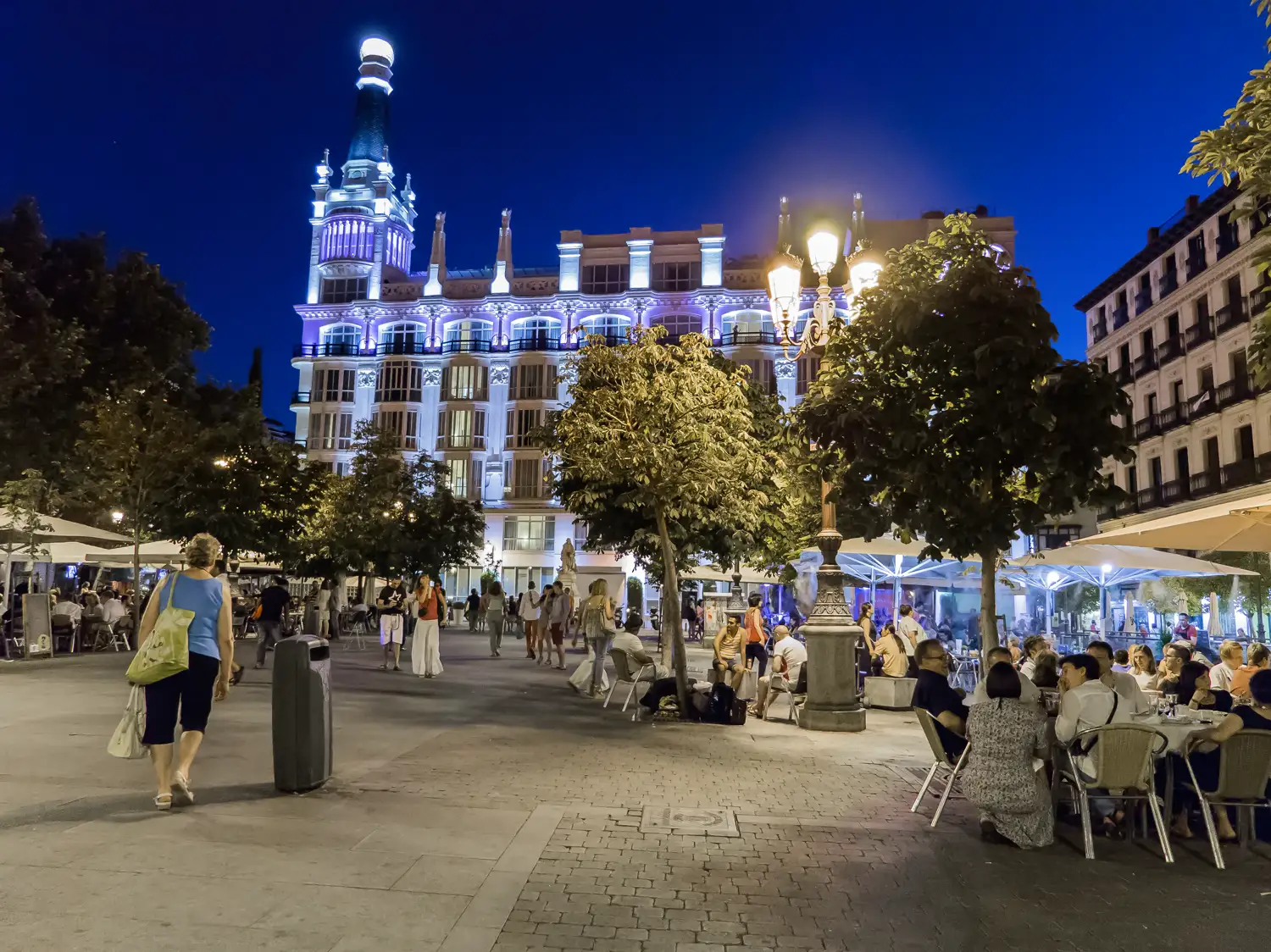 Terrazas llenas de gente sentada a mesas con sombrillas blancas en la plaza de Santa Ana de Madrid al anochecer, con el edificio iluminado del hotel ME Reina Victoria al fondo y viandantes cruzando la plaza