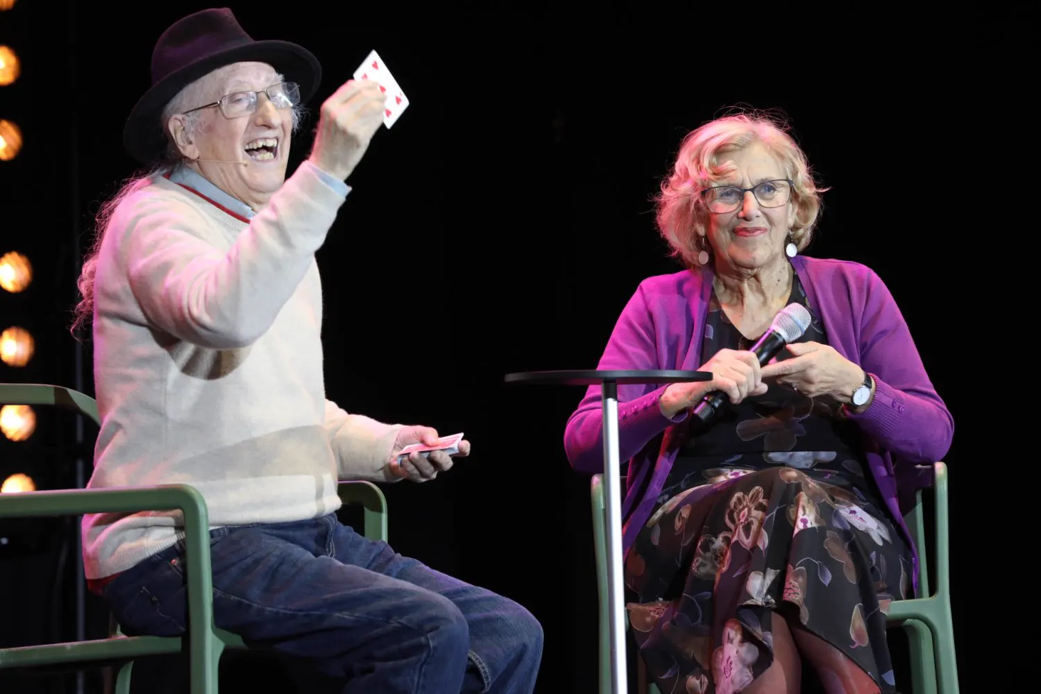Juan Tamariz, con chistera y gafas, ríe mientras muestra una carta sentado junto a Manuela Carmena en el escenario del Teatro Circo Price