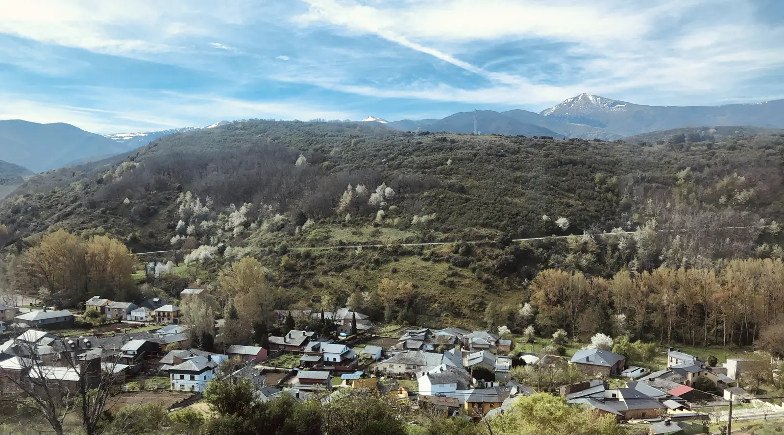 Vista desde lo alto de un pueblo de casas con tejado de pizarra en el fondo de un valle estrecho; alrededor suben laderas de monte con arbolado y, al fondo, cumbres con nieve bajo un cielo con nubes.