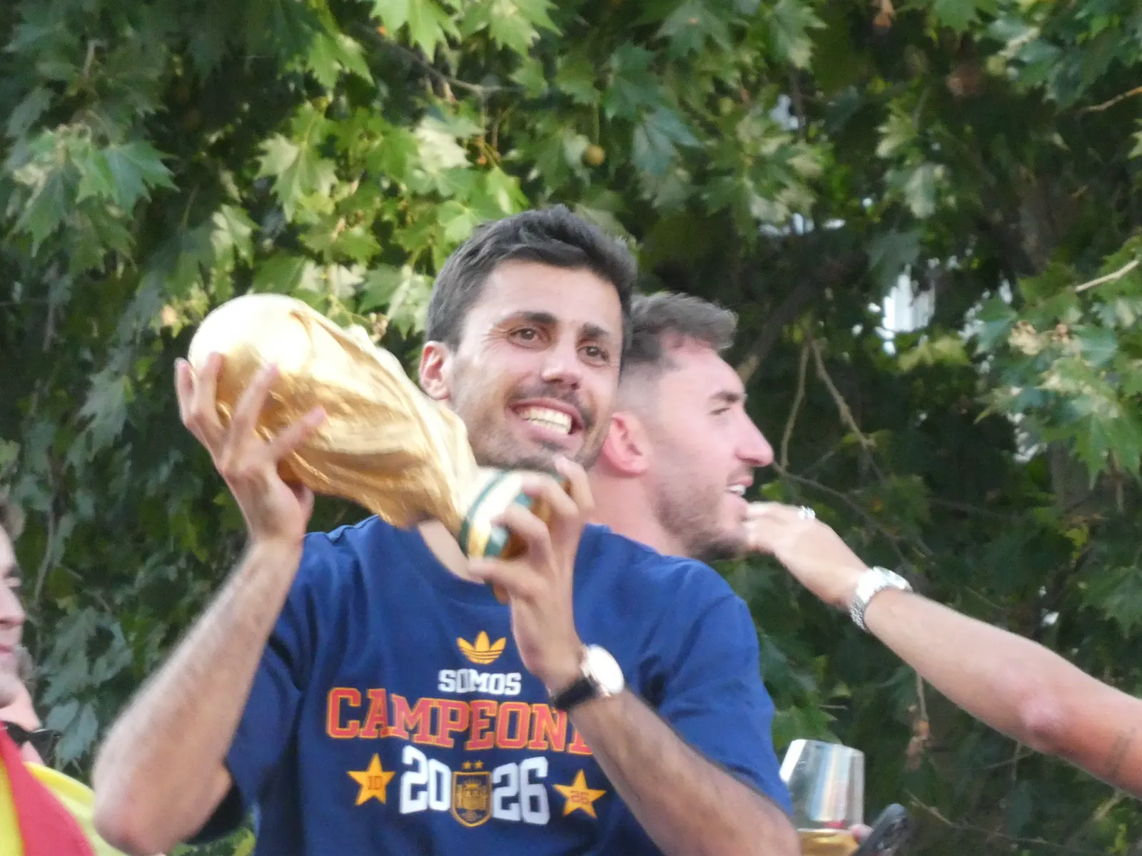 Rodri Hernández, con camiseta azul de la selección española que dice «Somos campeones 2026», levanta con las dos manos la Copa del Mundo dorada durante la celebración en un autobús descubierto