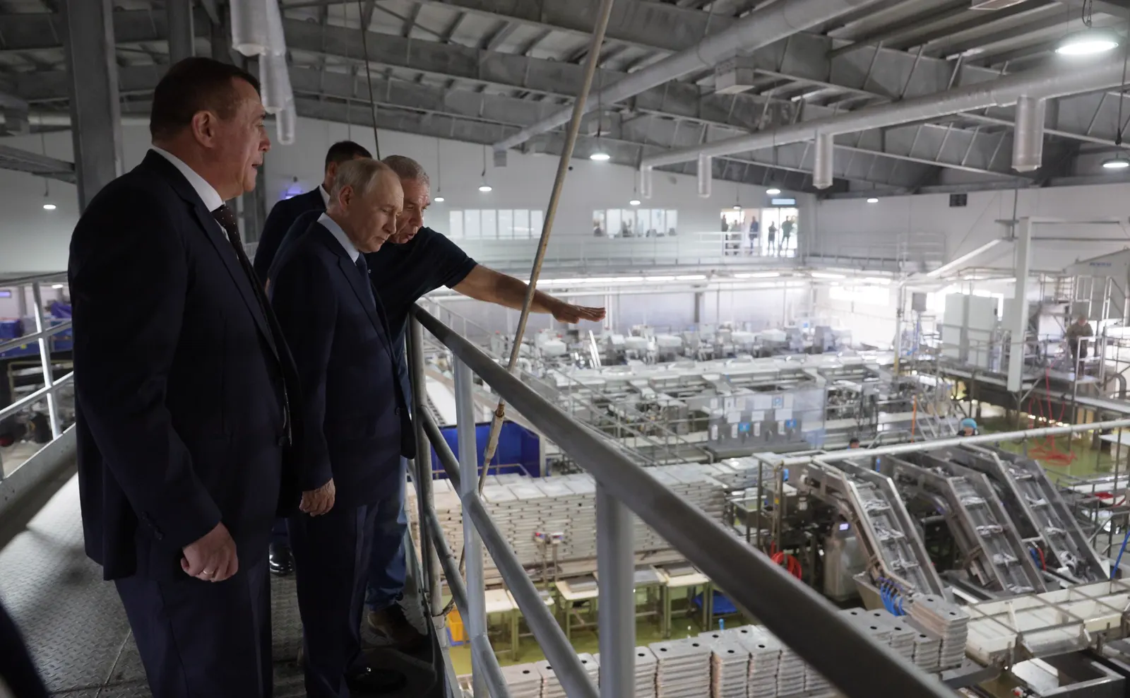 Vladimir Putin, con traje oscuro, observa desde una pasarela metálica la nave de una fábrica de procesado de pescado; a su lado, tres hombres, uno de ellos señalando con el brazo las líneas de producción.