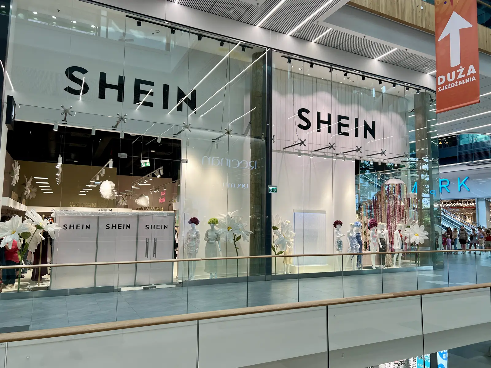 Escaparate acristalado de una tienda Shein en un centro comercial, con el rótulo de la marca y maniquíes entre flores blancas gigantes.