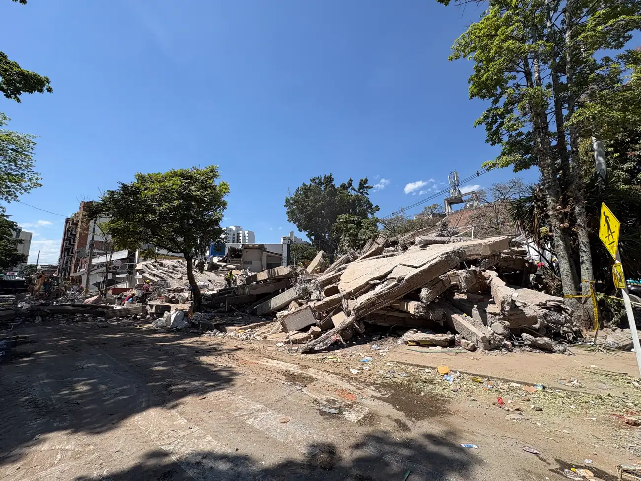 Solar despejado en una calle de Cali con una gran montaña de losas de hormigón amontonadas, resto de un edificio derrumbado. Al fondo, a la izquierda, un inmueble de varias plantas con la fachada abierta y operarios con casco trabajando entre los escombros; a la derecha, árboles, una señal amarilla de paso de peatones y cinta de balizamiento.