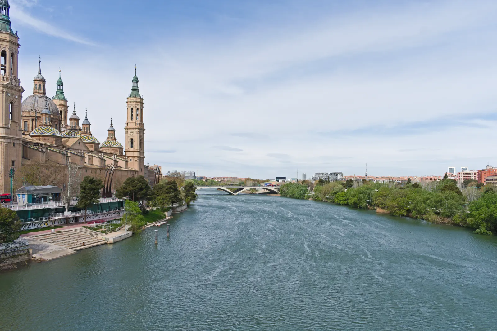 El río Ebro a su paso por Zaragoza visto desde un puente, con la basílica del Pilar y sus torres y cúpulas de tejas de colores alzándose sobre la ribera izquierda, arboledas en las dos orillas y un cielo azul con nubes altas.