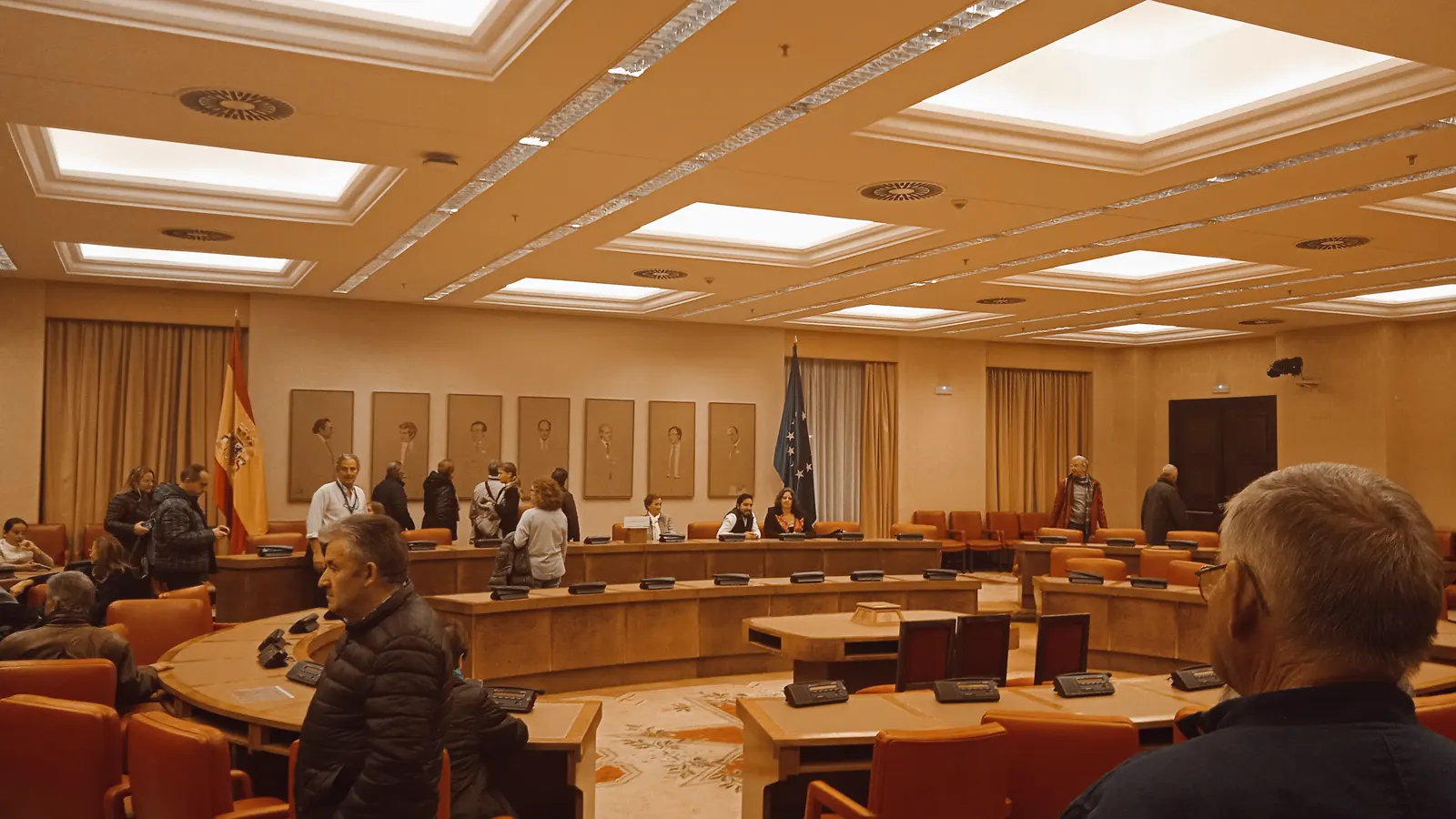 Sala Constitucional del Congreso de los Diputados: mesas de madera en herradura con micrófonos y pantallas, sillones tapizados de rojo, la bandera de España a la izquierda y la de la Unión Europea a la derecha, retratos alineados en la pared del fondo y techo artesonado con lucernarios. Varias personas recorren la sala de pie.