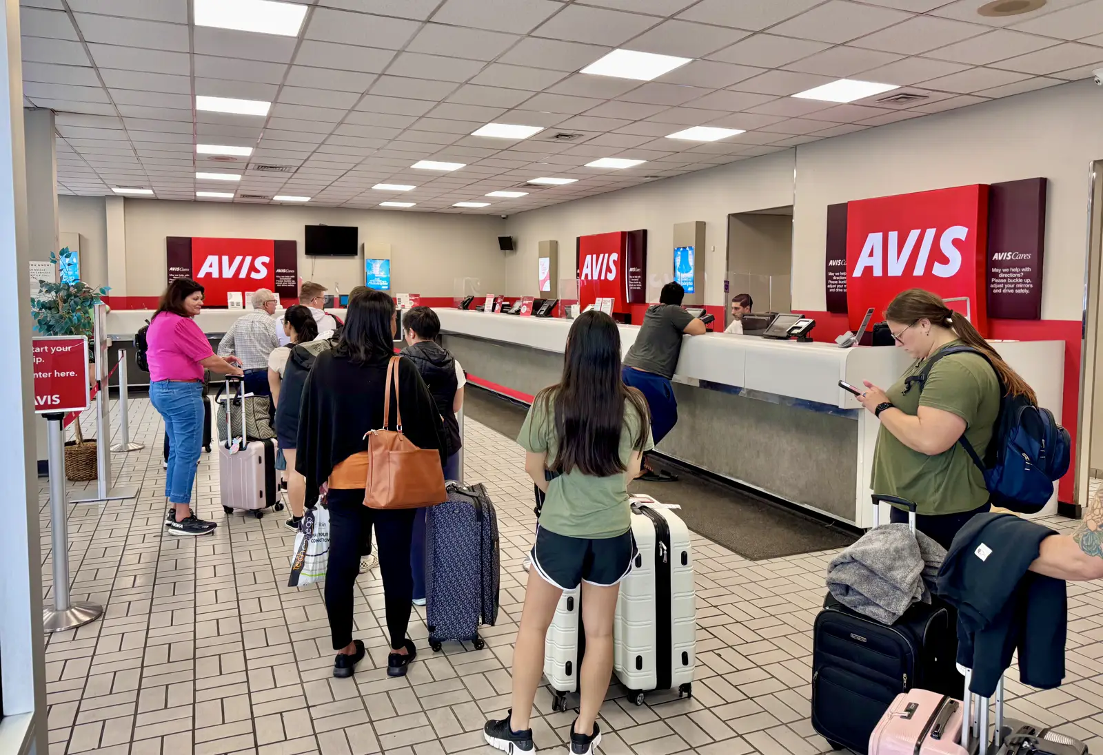 Cola de clientes con maletas ante los mostradores de alquiler de coches de Avis, con grandes rótulos rojos de la marca al fondo, en una terminal de aeropuerto.