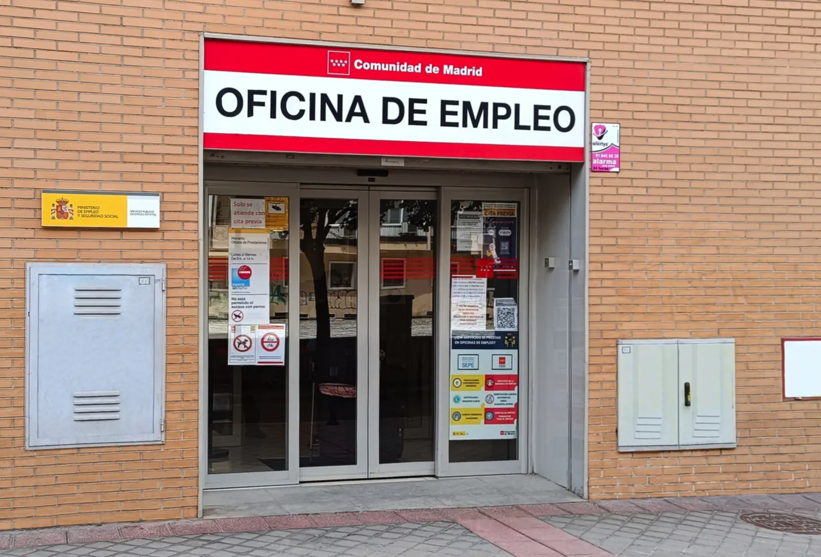 Entrada acristalada de una oficina de empleo con rótulo rojo de la Comunidad de Madrid y placa del Ministerio de Empleo y Seguridad Social - Servicio Público de Empleo Estatal en la fachada de ladrillo