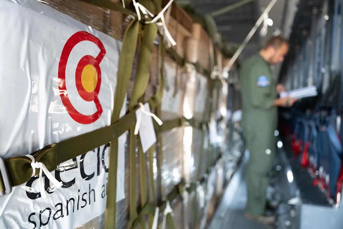 Palés de ayuda humanitaria envueltos en plástico con el logotipo de la AECID, sujetos con cinchas en la bodega de un avión; al fondo, un militar de espaldas revisa unos documentos.