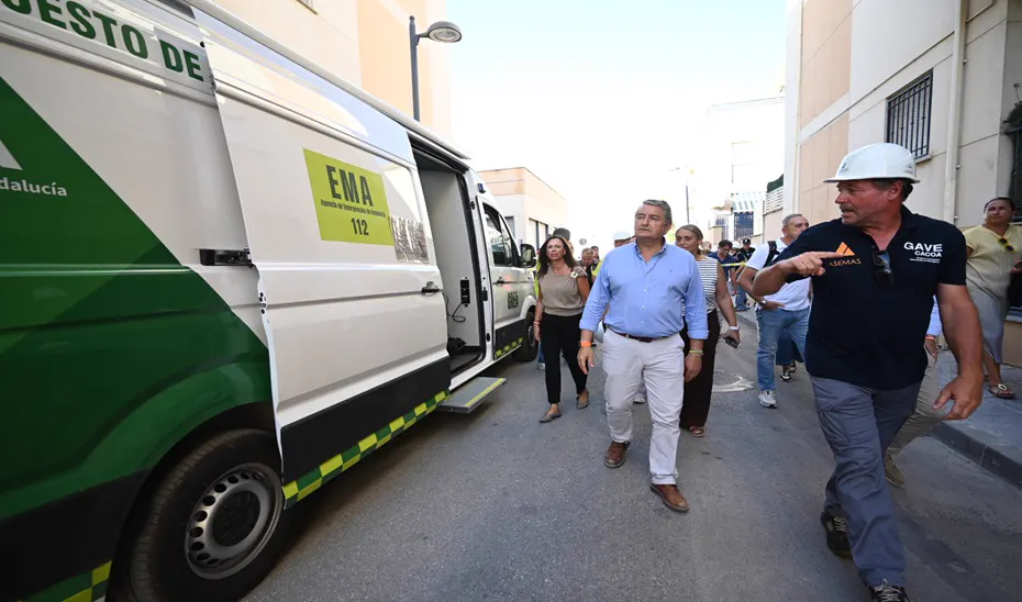 El consejero Antonio Sanz camina por una calle estrecha junto a la furgoneta blanca y verde del puesto de mando de la Agencia de Emergencias de Andalucía (EMA 1-1-2), con la puerta lateral abierta; a su lado, un voluntario con casco blanco y camiseta con el rótulo GAVE señala hacia las fachadas, y detrás caminan varias personas más.