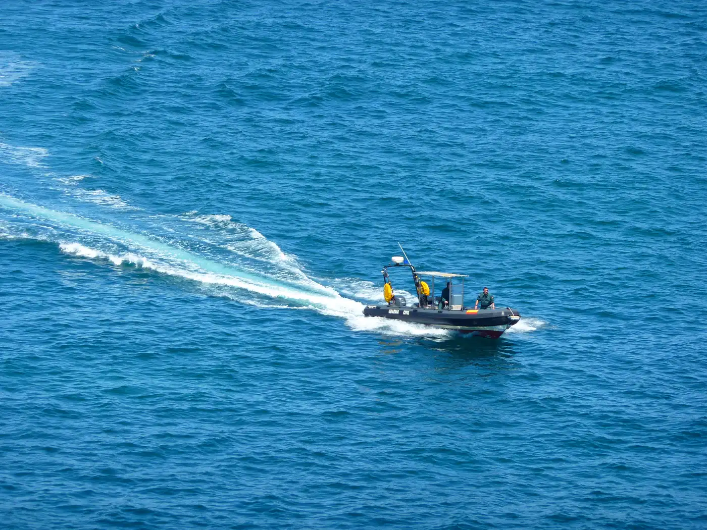 Lancha semirrígida oscura con el rótulo «Guardia Civil» navegando a velocidad en mar abierto, con dos tripulantes a bordo y una larga estela blanca a popa.