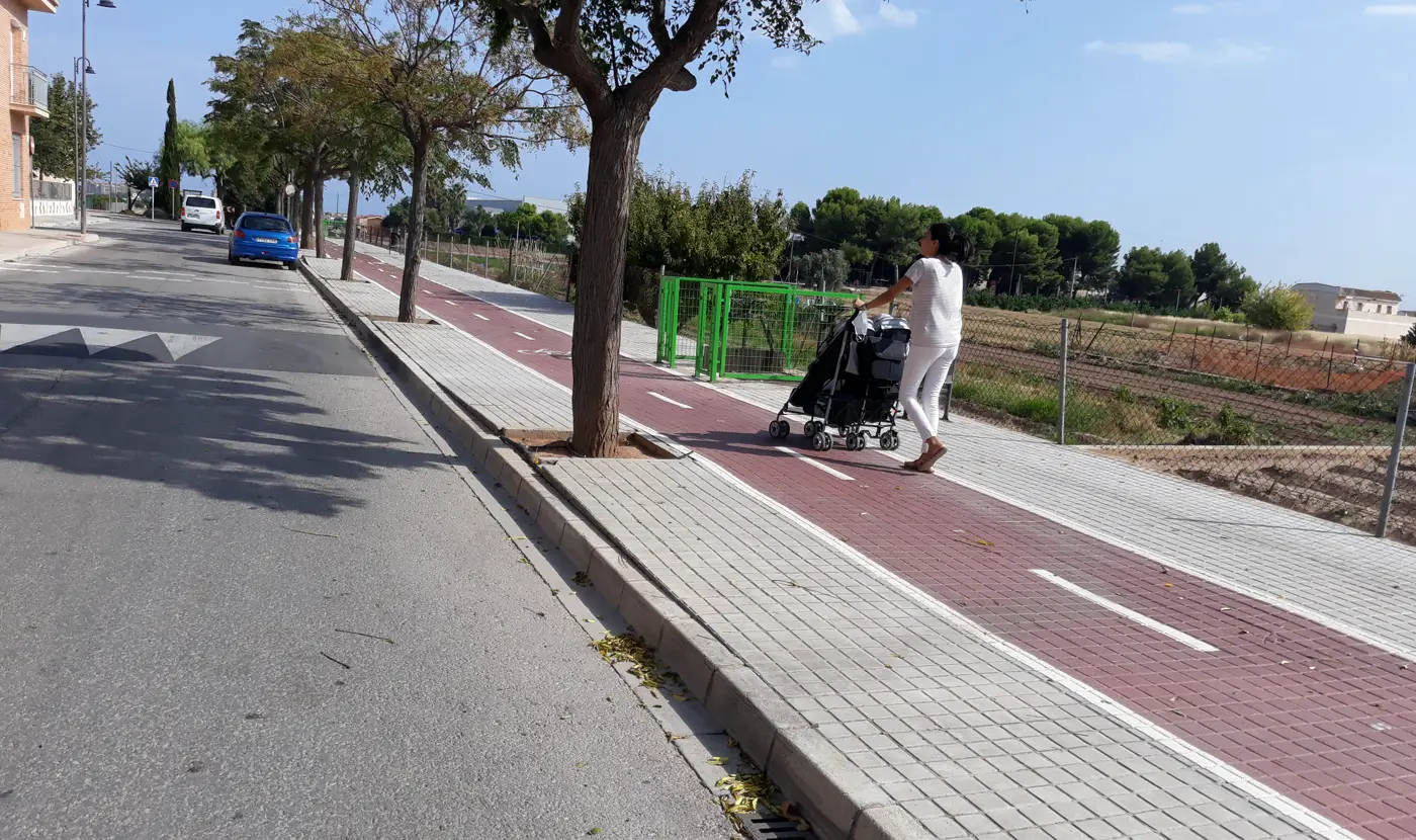 Una mujer vestida de blanco, de espaldas, empuja un cochecito doble de bebé por un paseo peatonal junto a un carril bici de asfalto rojo, con árboles a un lado y campos de cultivo al otro.