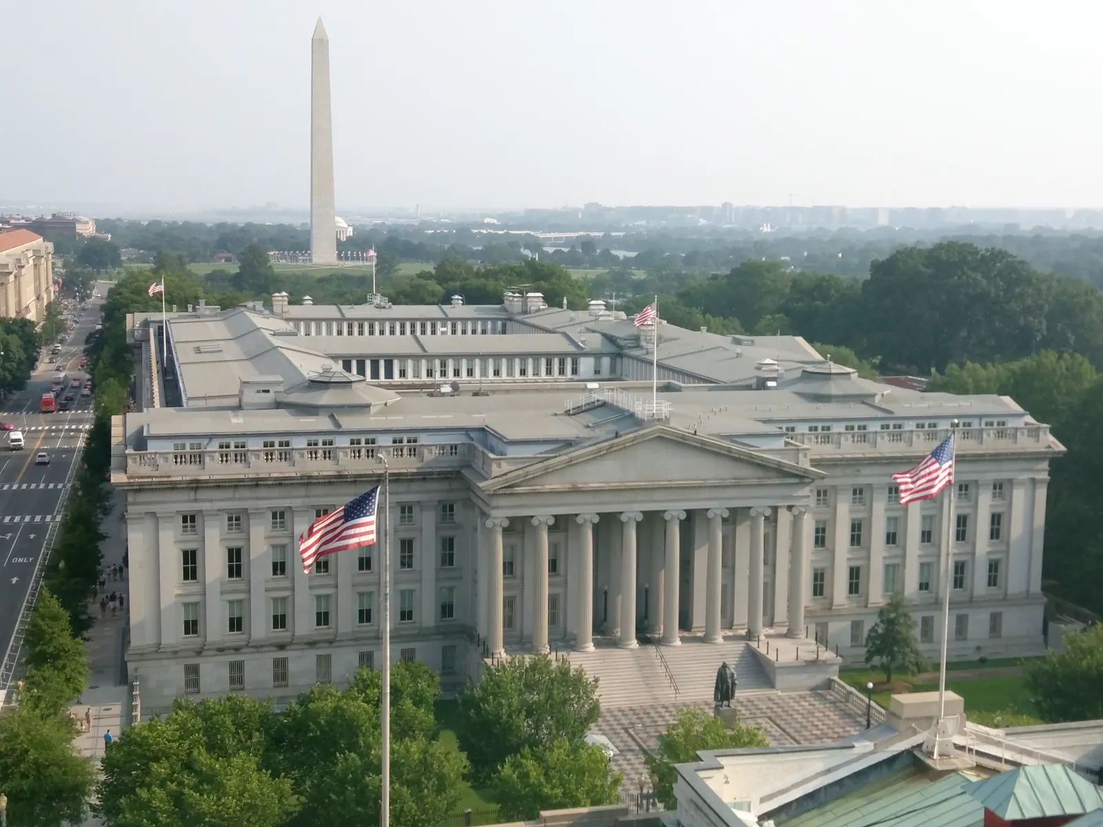 Vista aérea del edificio neoclásico del Departamento del Tesoro de Estados Unidos en Washington, con dos banderas estadounidenses en primer plano y el obelisco del Monumento a Washington al fondo.
