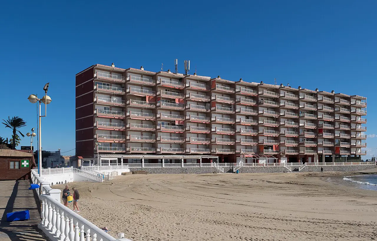 Bloque de apartamentos de siete alturas con balcones corridos y toldos rojizos, levantado justo al borde de la arena de una playa vacía, bajo un cielo azul sin nubes. En primer plano, una balaustrada blanca de paseo marítimo por la que bajan dos personas.