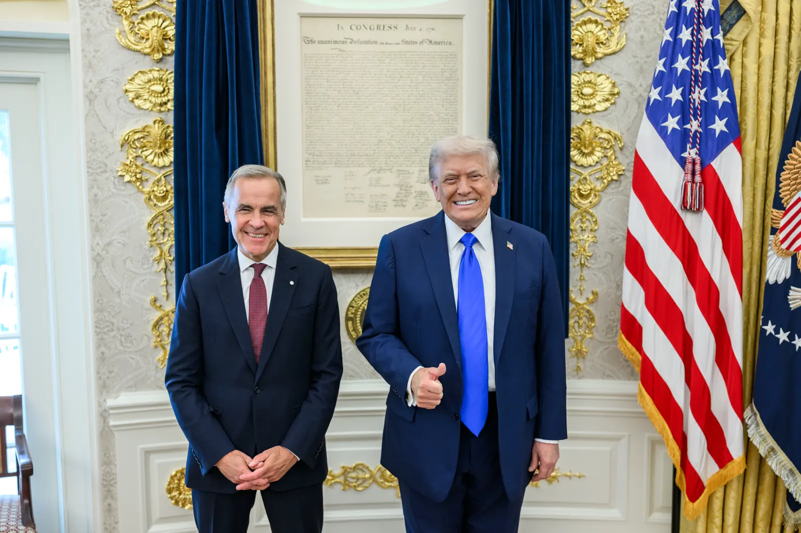 Mark Carney y Donald Trump, de pie y sonrientes en el Despacho Oval, ante una copia enmarcada de la Declaración de Independencia y la bandera de Estados Unidos; Trump levanta el pulgar.
