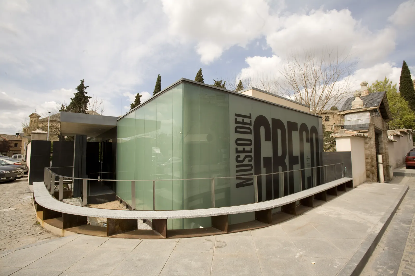 Pabellón acristalado de planta baja con el rótulo «Museo del Greco» en grandes letras negras sobre el vidrio verdoso, junto a una tapia de ladrillo y cipreses, bajo un cielo con nubes.