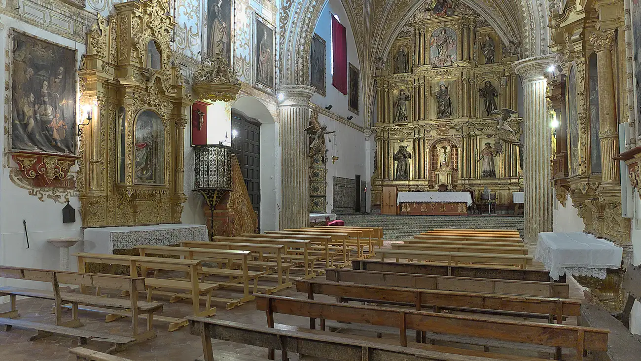 Interior de una iglesia conventual con retablos dorados a ambos lados, bancos de madera en la nave y el altar mayor al fondo bajo un arco apuntado.