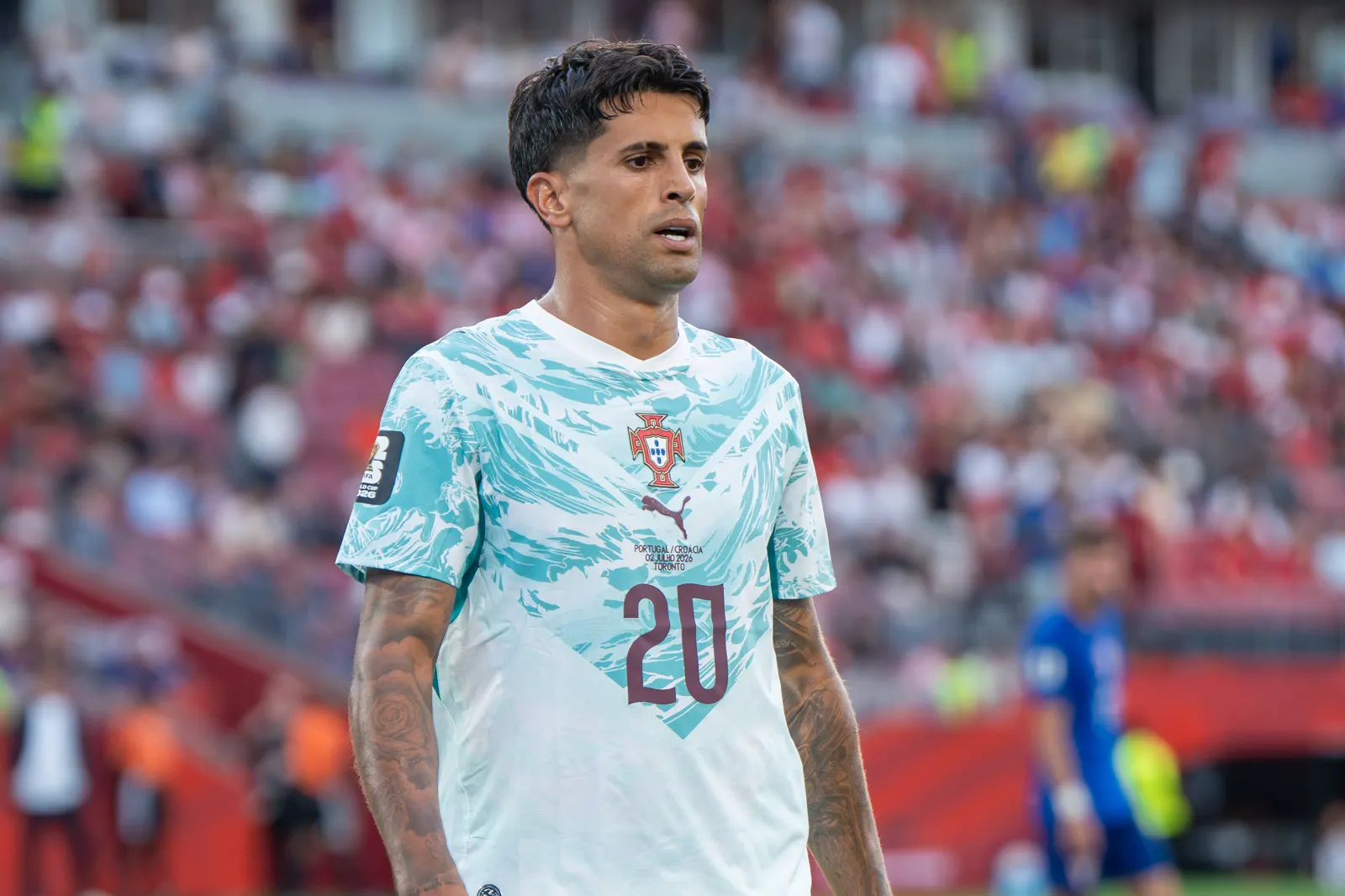 Primer plano de Joao Cancelo con la camiseta blanca y verde de la selección de Portugal, dorsal 20, sobre el césped y con la grada desenfocada al fondo