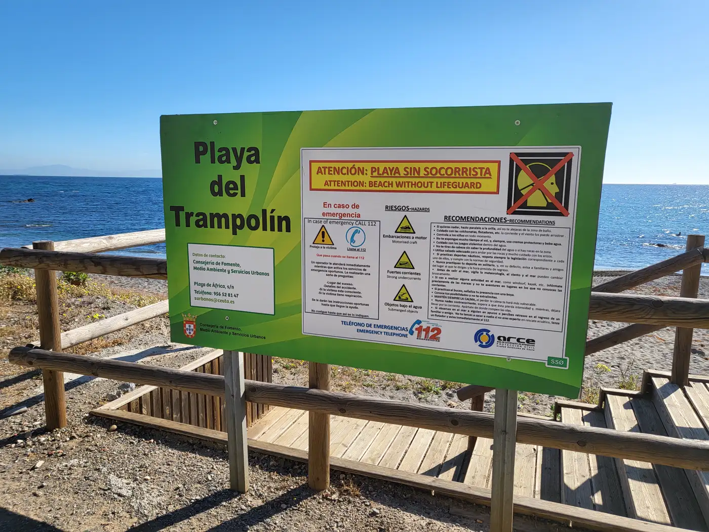 Cartel municipal verde con el rótulo «Playa del Trampolín» y un panel de recomendaciones de seguridad que avisa de que la playa no tiene socorrista, montado sobre postes junto a una valla y una pasarela de madera; al fondo, la orilla rocosa, el mar y el cielo despejado.