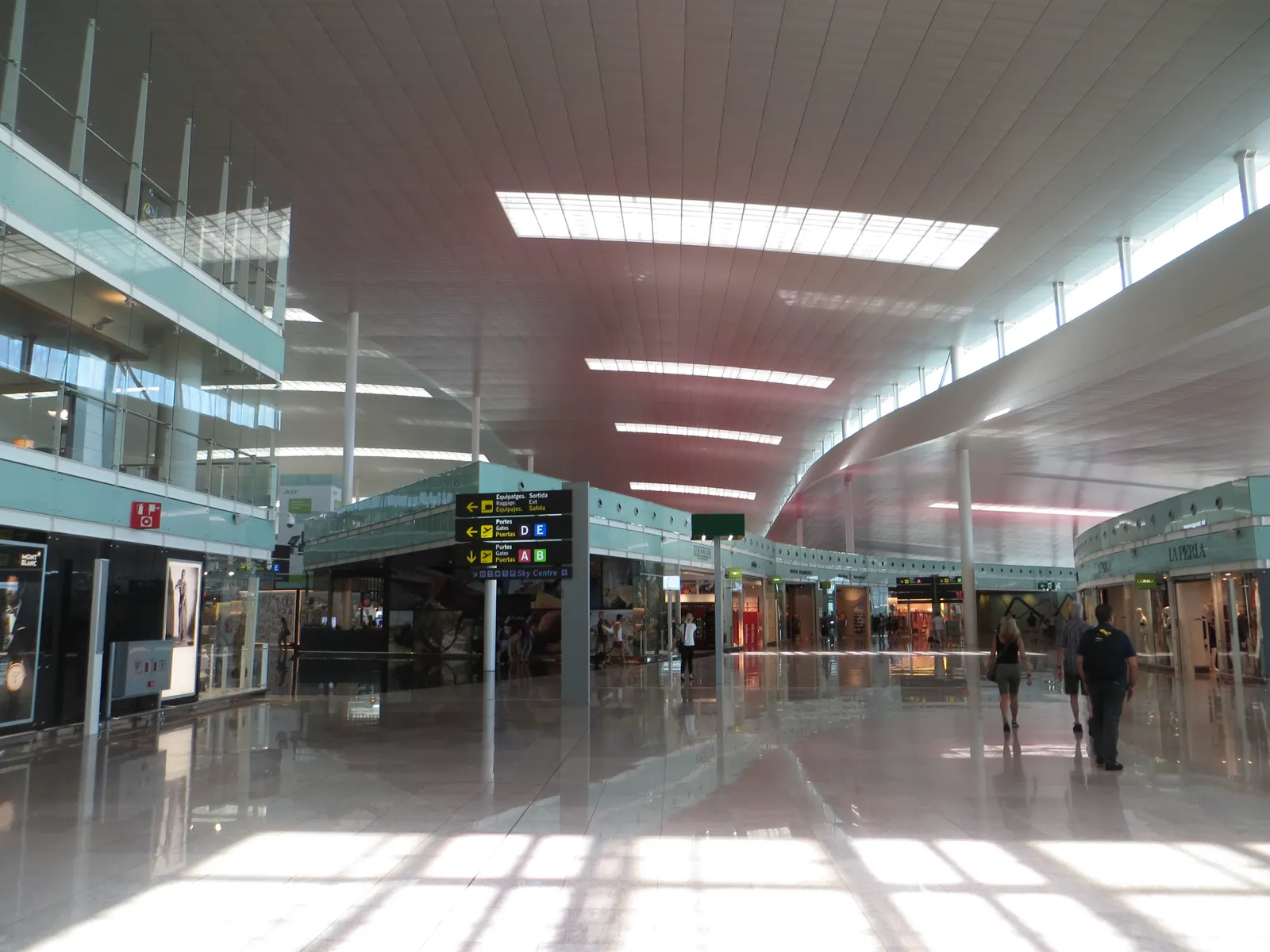 Vestíbulo de la Terminal 1 del aeropuerto de Barcelona-El Prat, con techo blanco ondulado, suelo pulido que refleja la luz, tiendas a ambos lados y un panel indicador de puertas de embarque; unos pocos viajeros caminan al fondo.