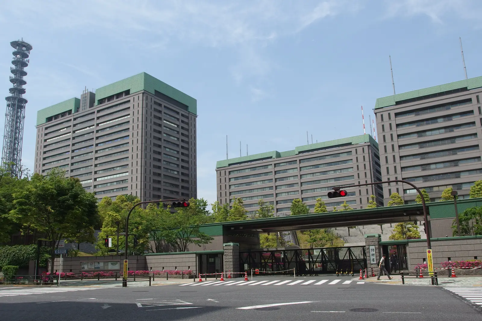 Complejo de edificios de oficinas de hormigón gris con remates verdes y una torre de antenas a la izquierda, vistos desde un cruce de calles vacío con semáforos en rojo, un paso de cebra y una puerta de acceso vigilada al fondo.