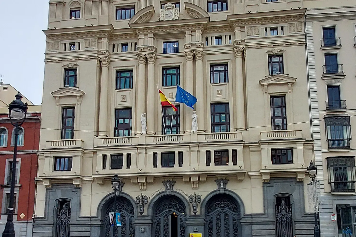 Fachada de piedra clara del Ministerio de Educación en la calle Alcalá 34 de Madrid, con columnas, la bandera de España y la de la Unión Europea ondeando sobre el balcón principal y tres portones de hierro forjado en la planta baja.