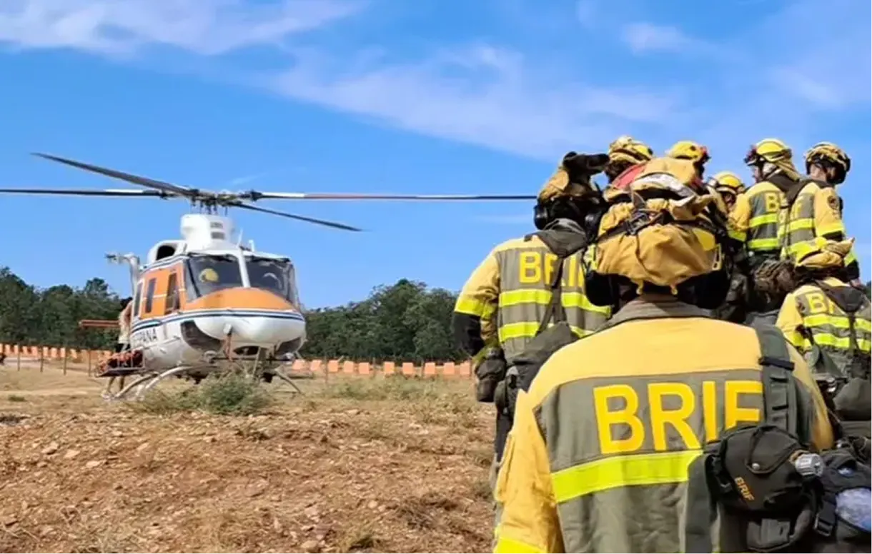 Una decena de bomberos forestales con uniforme amarillo y gris, mochilas y cascos, vistos de espaldas mientras caminan por un rastrojo seco hacia un helicóptero naranja y blanco con las palas en marcha. En la espalda de los monos se lee BRIF. Al fondo, una línea de arbolado verde y cielo azul.