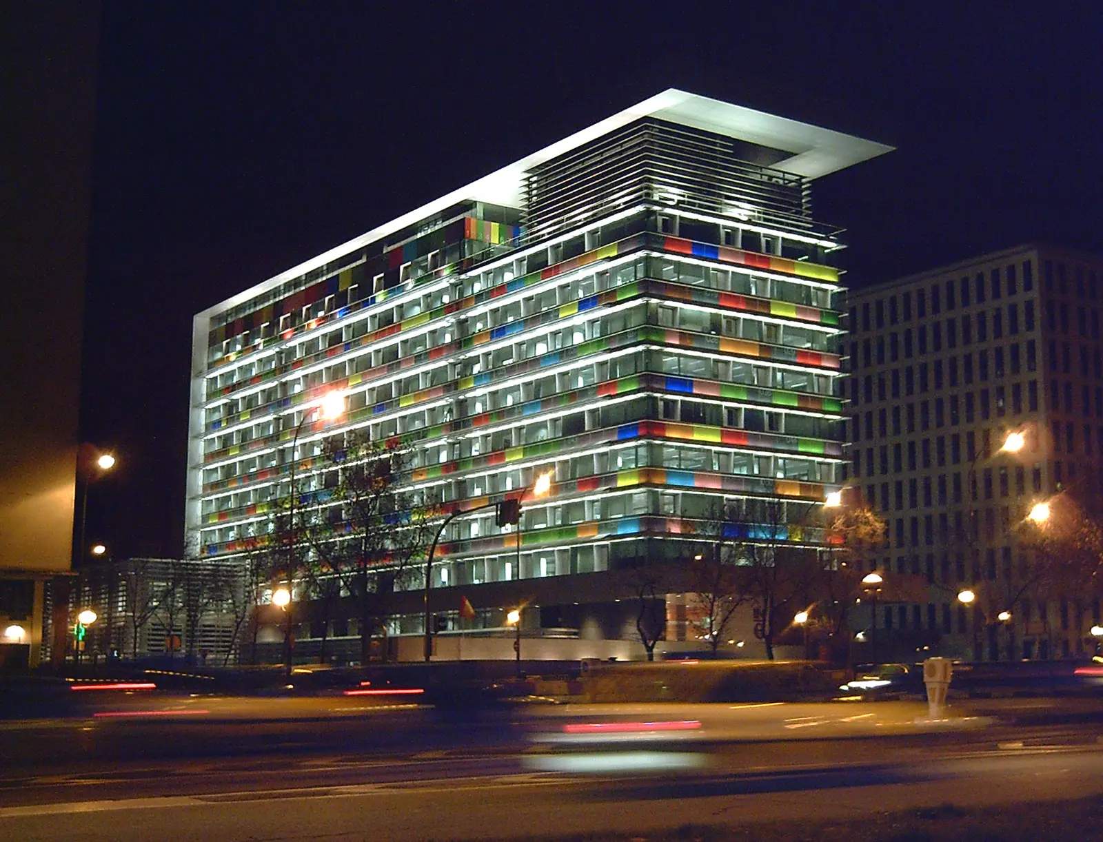 Vista nocturna de un edificio de oficinas de una decena de plantas con la fachada acristalada iluminada y paneles de colores rojos, verdes, azules y amarillos intercalados entre las ventanas, junto a una avenida por la que pasan coches con las luces trazadas.