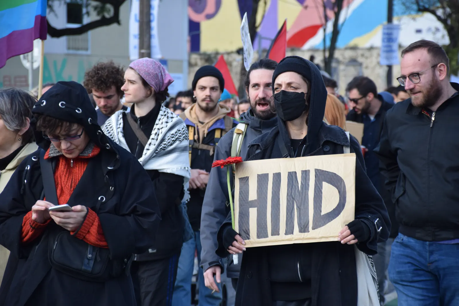 Manifestación en una calle de Lisboa: una mujer con capucha y mascarilla negras sostiene un cartel de cartón con la palabra HIND escrita a mano, rodeada de otros manifestantes con banderas y pañuelos palestinos