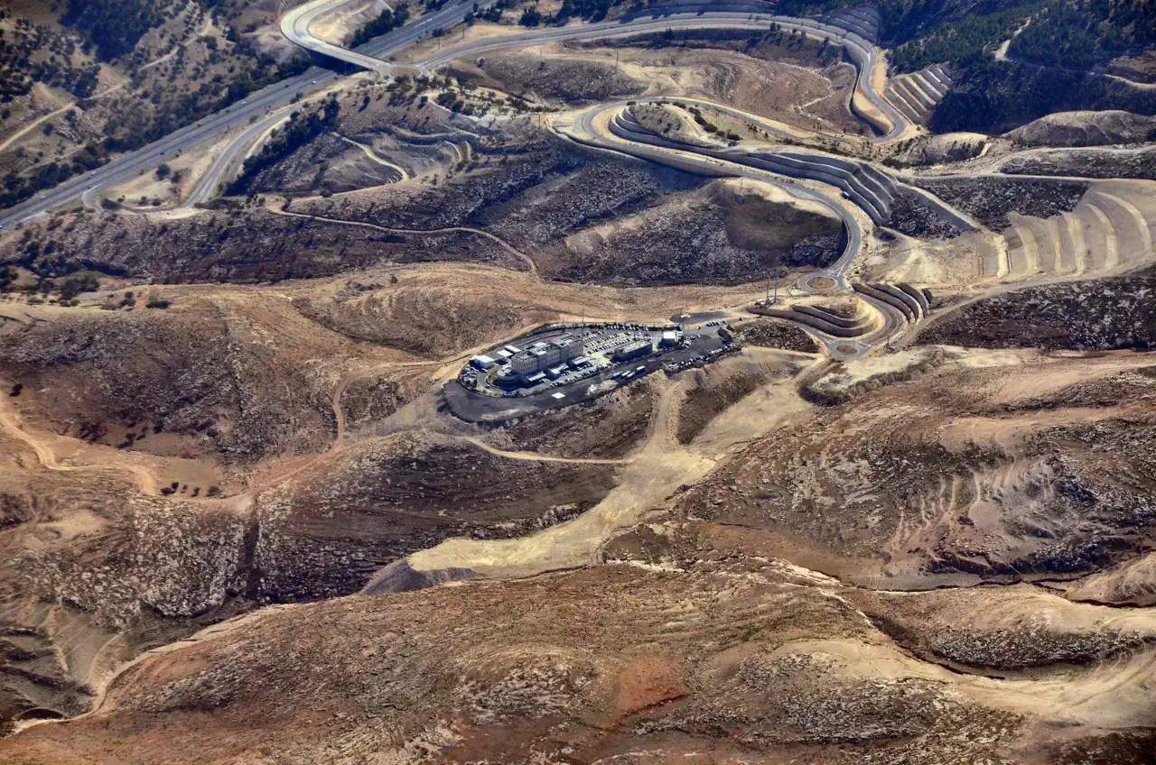 Vista aérea de colinas áridas y sin edificar del área E1, cruzadas por carreteras y por un enlace de autopista, con un recinto vallado de edificios bajos y aparcamiento en el centro de la imagen.